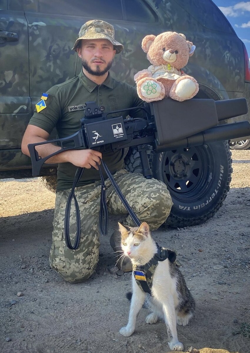 A soldier with an anti-drone gun with a teddy bear on it holds a cat on a leash.