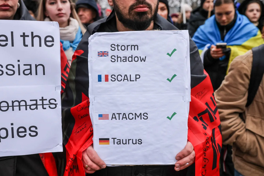 A protestor holds a sign where Storm Shadow, SCALP, and ATACMS are checked off, but Taurus is not.